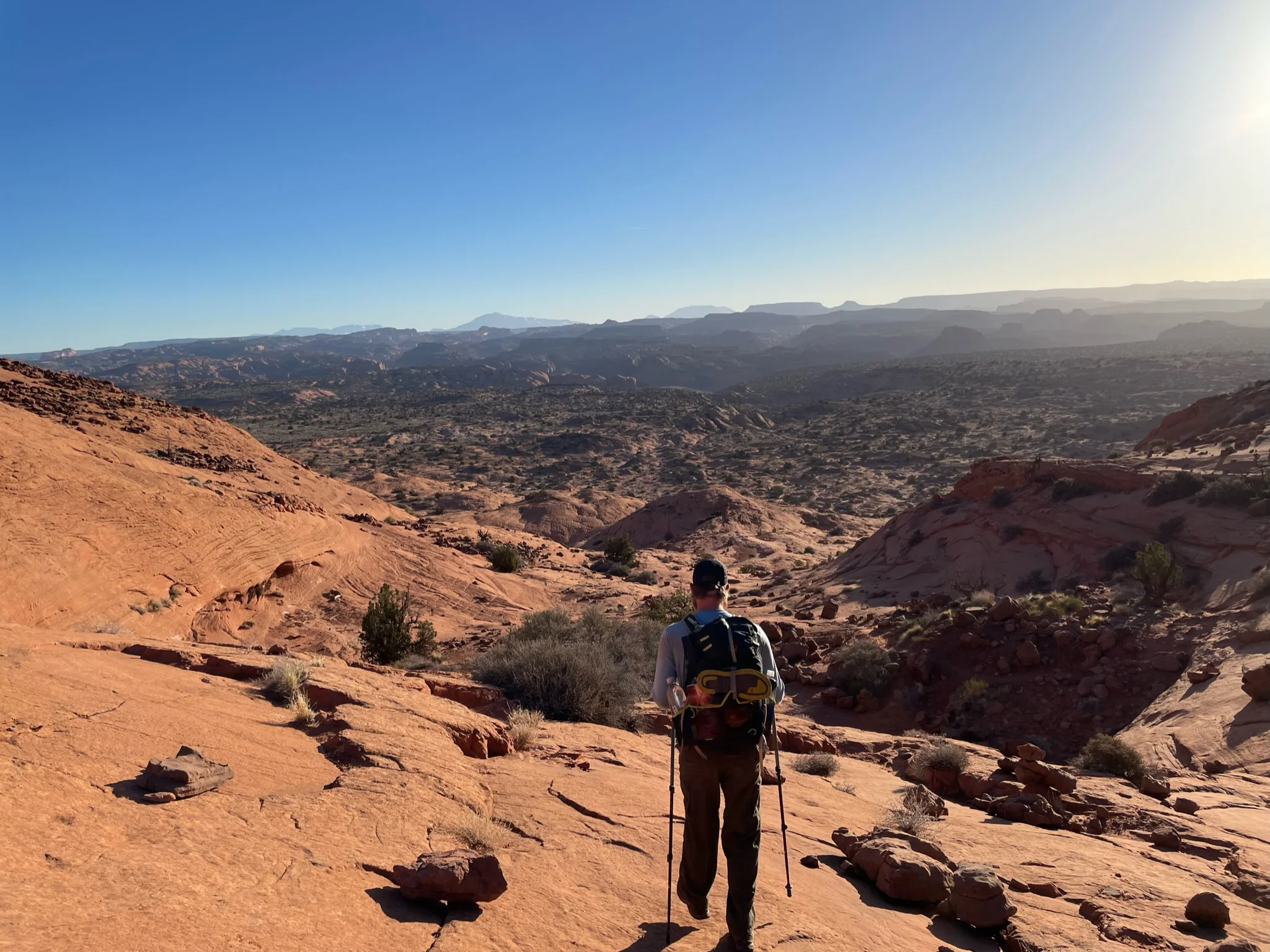 A backpacker with trekking poles walking across red sandstone above a desert valley.